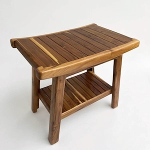 May include: A wooden shower stool with a slatted seat and a lower shelf. The stool is made of light and dark brown wood and has a curved seat design. The legs are square and sturdy. The stool is designed for use in a shower or bathroom.