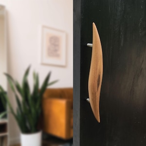 May include: A wooden door handle with a curved, leaf-like design. The handle is made of light brown wood and is attached to a black door with two silver metal screws. The background is a blurred interior.