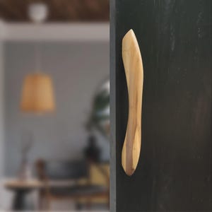 Könnte beinhalten: Ein hölzerner Türgriff mit geschwungenem, ergonomischem Design. Der Griff besteht aus hellem Holz mit dunkleren Maserungsakzenten. Er ist an einer dunklen, möglicherweise schwarzen Tür oder Oberfläche montiert.