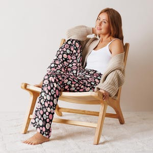 Könnte beinhalten: Eine Frau in einer schwarzen Hose mit rosafarbenem Muster sitzt auf einem Holzstuhl. Die Hose hat ein großes Kreis-Muster mit rosa, weißen und grauen Kreisen auf schwarzem Hintergrund.