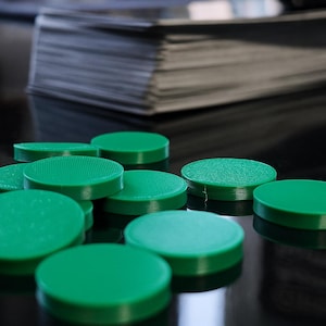 Könnte beinhalten: Helle, grüne, kreisförmige Spielchips liegen auf einer reflektierenden, schwarzen Oberfläche. Einige Chips haben eine strukturierte Oberfläche. Kartenstapel im Hintergrund deuten auf ein Brett- oder Kartenspiel hin.