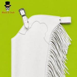 May include: A pair of white leather chaps featuring long fringe along the side, a scalloped yoke design, and an adjustable belt with a silver-toned metal buckle. The item is displayed against a bright lime green background with a skull logo.