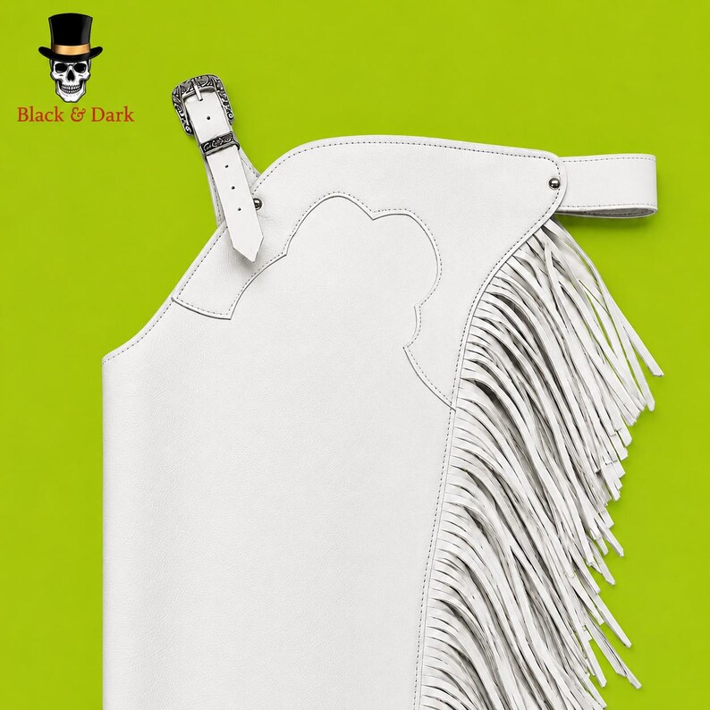 May include: A pair of white leather chaps featuring long fringe along the side, a scalloped yoke design, and an adjustable belt with a silver-toned metal buckle. The item is displayed against a bright lime green background with a skull logo.