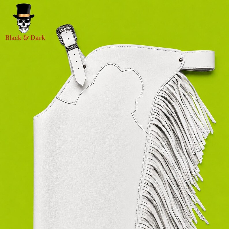 May include: A white leather chaps garment featuring long fringe detailing along the side, a decorative silver-toned metal buckle closure at the top, and a wide adjustable waist strap. The design includes stitched paneling and a smooth finish.