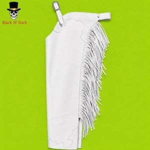 May include: A white leather chap featuring long fringe detailing along the side, a scalloped yoke design, and an adjustable belt strap with a silver-toned metal buckle. The item is displayed against a bright lime green background.