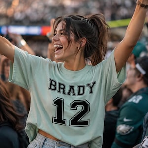 May include: A light mint green short-sleeved t-shirt featuring the name BRADY in black block letters above the number 12 in a large black font. The shirt is made of cotton and has a crew neckline, worn with light blue denim jeans.