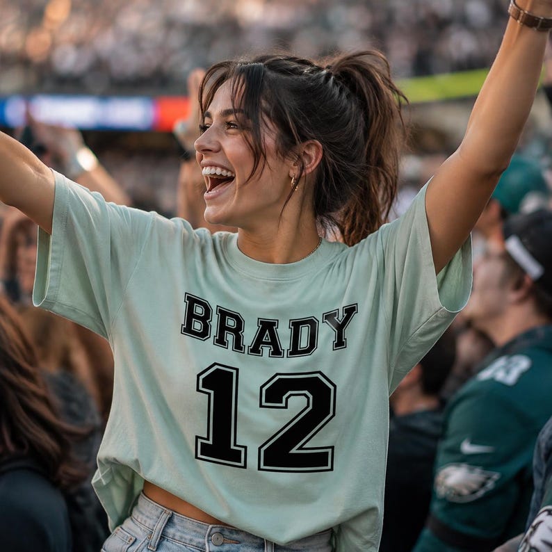 May include: A light mint green short-sleeved t-shirt featuring the name BRADY in black block letters above the number 12 in a large black font. The shirt is made of cotton and has a crew neckline, worn with light blue denim jeans.