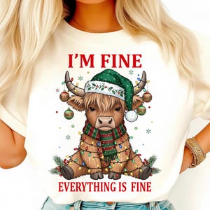 Könnte beinhalten: Cremefarbenes Sweatshirt mit einer Illustration einer Highland-Kuh. Die Kuh trägt einen grünen Weihnachtsmannhut, einen rot-grünen Schal und ist mit Lichterketten und Weihnachtskugeln geschmückt. Der Text "I'M FINE EVERYTHING IS FINE" ist über und unter der Kuh gedruckt.