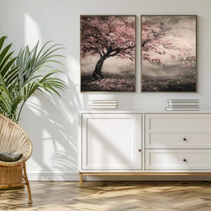 a serene indoor setting with a large tree in full bloom, its branches adorned with pink flowers, casting a shadow on the wall. Above the tree, two framed paintings are displayed, one depicting a tree with pink flowers and the other showing a landscape with a river and mountains. The paintings are hung on a white wall, and a wicker chair is positioned in the foreground, adding to the cozy and inviting atmosphere of the room.