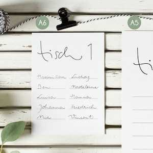 Puede incluir: Se muestran dos tarjetas blancas con el texto "tisch 1" en escritura negra. Una tarjeta tiene una lista de nombres, la otra tiene líneas para nombres. Las tarjetas están sujetas a una cuerda sobre un fondo de madera blanca.