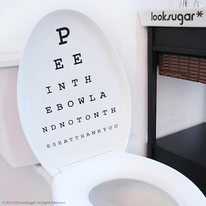 Puede incluir: Asiento de inodoro blanco con una calcomanía de tabla de agudeza visual negra que dice "P EE INTH EBOWLA NDNOTONTH ESEATTHANKYOU".