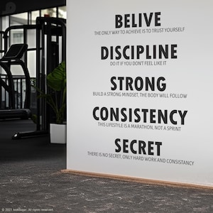 May include: A white wall in a gym setting features motivational text in black font: BELIVE, DISCIPLINE, STRONG, CONSISTENCY, and SECRET. The text is accompanied by supporting phrases. Gym equipment is visible in the background.