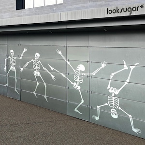 May include: A gray garage door features a playful design of white skeleton figures in various dancing poses. The skeletons are depicted with outstretched arms and legs, creating a dynamic and whimsical scene. The text "looksugar*" is visible at the top.