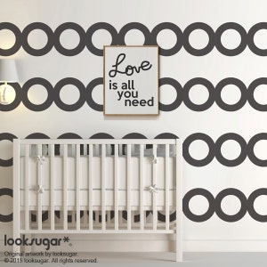 Peut inclure: Un lit bébé blanc avec un matelas et des oreillers gris est placé devant un mur décoré d'un motif répétitif de cercles gris foncé imbriqués. Une affiche encadrée avec les mots "Love is all you need" est accrochée au mur.