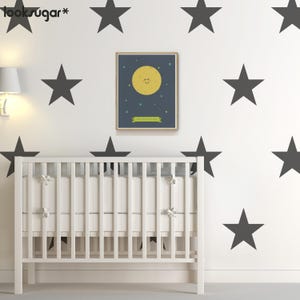 May include: A white crib with a white mattress and a white wooden frame. The crib is in front of a white wall with black star stickers. A framed print of a yellow moon with a smiling face is hanging on the wall.
