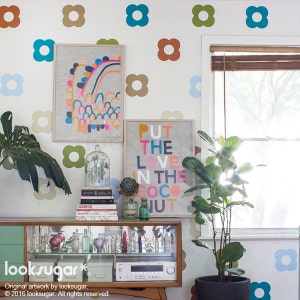 May include: A room decorated with framed artwork, a cabinet, and a potted plant. The artwork includes a colorful abstract design and a print that says "PUT THE LOVE IN THE COCONUT." The wall has flower-shaped decals.