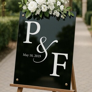 Puede incluir: Un letrero de boda negro con letras blancas que muestra las iniciales "P & F" y la fecha "18 de mayo de 2019". El letrero est&aacute; adornado con flores blancas y vegetaci&oacute;n en la parte superior y est&aacute; montado en un caballete de madera.