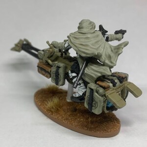 Może przedstawiać: Miniaturowa figurka zamaskowanego jeźdźca na futurystycznym motocyklu. Jeździec ma na sobie jasny zielony płaszcz, a motocykl jest oliwkowozielony z brązowymi akcentami. Podstawa to brązowy owal z kępką suchej trawy.