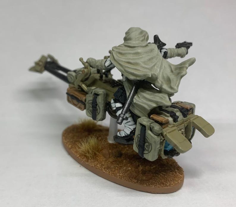 Może przedstawiać: Miniaturowa figurka zamaskowanego jeźdźca na futurystycznym motocyklu. Jeździec ma na sobie jasny zielony płaszcz, a motocykl jest oliwkowozielony z brązowymi akcentami. Podstawa to brązowy owal z kępką suchej trawy.