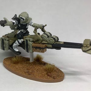 Może przedstawiać: Miniaturowa figurka futurystycznego żołnierza na poduszkowcu. Żołnierz ubrany jest w biało-czarny mundur z kapturem. Oliwkowy poduszkowiec ma brązowe i czarne detale i jest zamontowany na brązowej podstawie.