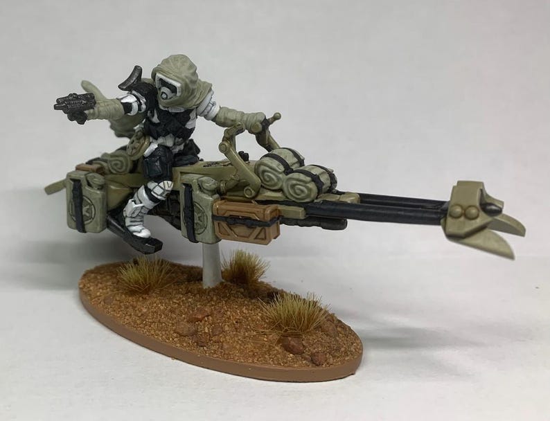 Może przedstawiać: Miniaturowa figurka futurystycznego żołnierza na poduszkowcu. Żołnierz ubrany jest w biało-czarny mundur z kapturem. Oliwkowy poduszkowiec ma brązowe i czarne detale i jest zamontowany na brązowej podstawie.