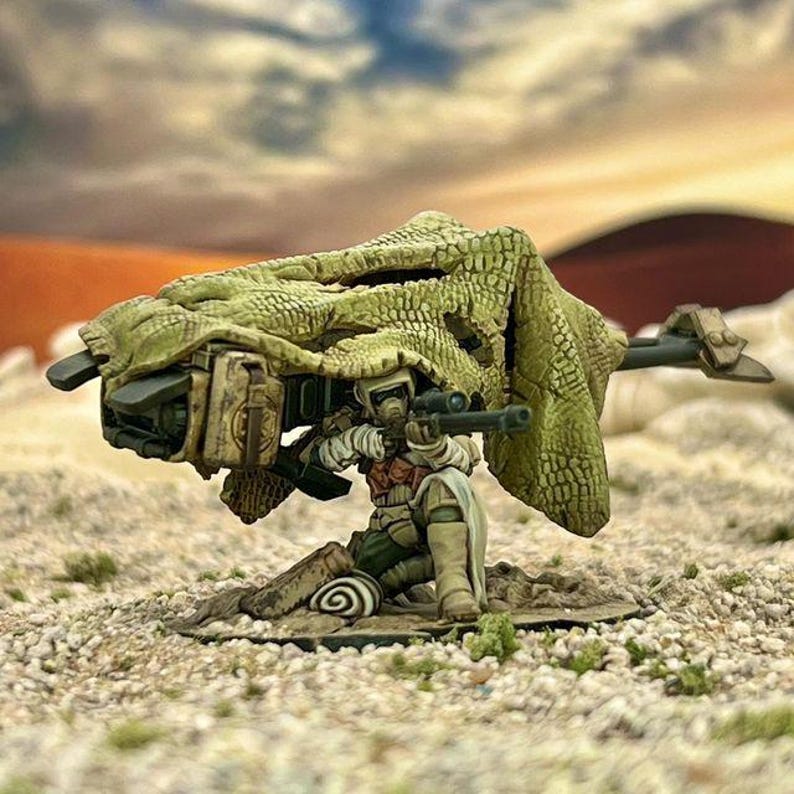 Może przedstawiać: Miniaturowa figurka żołnierza klęczącego z karabinem snajperskim, ustawiona za zamaskowanym działem artyleryjskim. Figurka jest pomalowana w stonowanych odcieniach, na tle pustynnego krajobrazu z zachodem słońca. Temat militarny lub fantasy.
