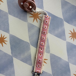 May include: A dusty pink dummy attached to a fabric strap featuring a repeating white and orange floral pattern. The strap is bordered by cream-coloured crochet trim and connects to a round, natural wood clip at the bottom via a metal fastener.