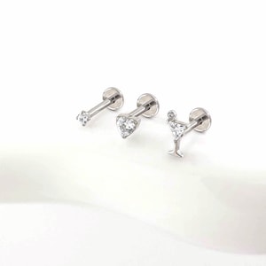 Può includere: Tre piercing per labbra argentati. Uno presenta una gemma trasparente e rotonda, un altro una gemma a forma di cuore e il terzo un design a bicchiere da martini con una piccola gemma. Tutti hanno un retro piatto e un perno.