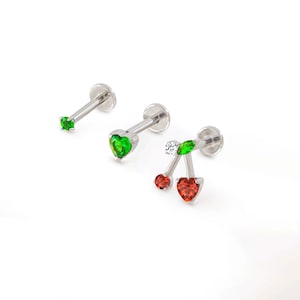 Könnte beinhalten: Drei silberfarbene Labret-Ohrstecker. Einer hat einen kleinen grünen Edelstein, ein anderer einen herzförmigen grünen Edelstein und der dritte ein Kirschdesign mit roten herzförmigen Edelsteinen, einem grünen Blatt und einem kleinen weißen Edelstein.