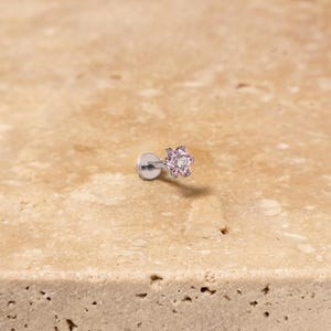Könnte beinhalten: Kleiner, silberfarbener Ohrring mit blütenförmigem Design. Die Blüte hat rosa Edelsteine um einen zentralen, klaren Edelstein. Für ein Knorpelpiercing.