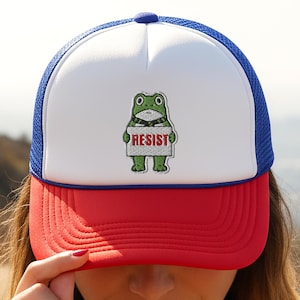 Peut inclure: Casquette de camionneur rouge, blanche et bleue avec un motif de grenouille tenant une pancarte avec le mot "RESIST". La casquette a un panneau avant blanc, une visière rouge et un dos en filet bleu.