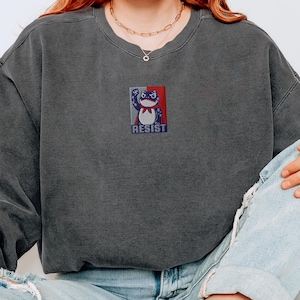 Peut inclure: Sweat-shirt gris foncé avec un petit dessin d'une grenouille levant le poing, avec le mot "RESIST" en dessous. La grenouille est rouge, blanche et bleue. Le sweat-shirt est de style col rond.