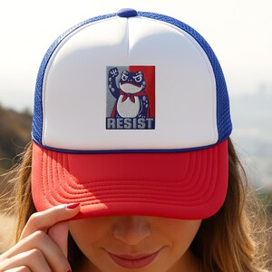 Peut inclure: Casquette de camionneur rouge, blanche et bleue avec un motif de grenouille. La grenouille est représentée avec un poing levé et une cape, avec le mot "RESIST" en dessous. La casquette a un panneau avant blanc, une visière rouge et un dos en filet bleu.