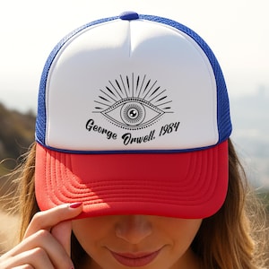 Peut inclure: Casquette de camionneur rouge, blanche et bleue avec le texte "George Orwell, 1984" et un graphisme d'œil. La casquette a un panneau avant blanc, une visière rouge et un dos en filet bleu. La casquette est portée.