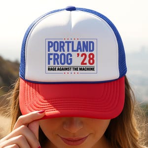 May include: A red, white, and blue trucker hat with the text "PORTLAND FROG '28 RAGE AGAINST THE MACHINE" in a blue rectangle. The hat has a white front panel, a red brim, and a blue mesh back.