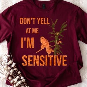 Puede incluir: Camiseta burdeos con el texto "DON'T YELL AT ME I'M SENSITIVE" en naranja. La camiseta presenta un gráfico de una planta y una pequeña criatura naranja. Un ramo de flores blancas está en la esquina inferior izquierda.