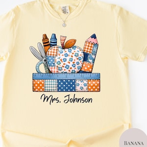 Può includere: T-shirt giallo chiaro con una grafica colorata di materiale scolastico, tra cui forbici, pastelli e una matita, disposti attorno a una mela patchwork. Il testo "Mrs. Johnson" è stampato sotto.
