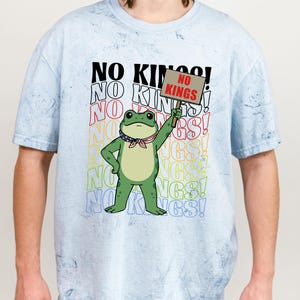 May include: Light blue t-shirt with a graphic of a green frog holding a sign that reads "NO KINGS!". The shirt also features the text "NO KINGS!" in a repeating pattern. The frog is wearing a red, white, and blue bandana.
