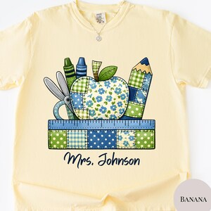 May include: A light yellow t-shirt featuring a colorful graphic of school supplies, including an apple, pencil, scissors, and ruler. The apple has a patchwork design with blue floral patterns. The text "Mrs. Johnson" is below the graphic.