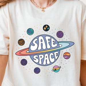 Puede incluir: Camiseta color crema con un diseño de planeta colorido con las palabras "SAFE SPACE" en una fuente de burbujas. El planeta tiene un anillo arcoíris y está rodeado de planetas más pequeños en varios colores y patrones.
