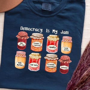 May include: A navy blue t-shirt with the slogan "Democracy Is My Jam." The design includes illustrations of jam jars, each with a label such as "Equality" and "Inclusion."