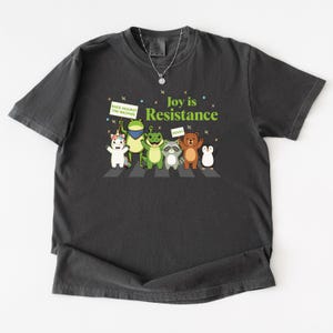 May include: Dark gray t-shirt featuring a whimsical illustration of various animals crossing a crosswalk. The text "Joy is Resistance" is above the animals. One animal holds a sign that says "Rage Against The Machine", and another holds a sign that says "Resist".