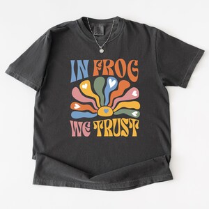 Może przedstawiać: Ciemnoszara koszulka z napisem "IN FROG WE TRUST" w stylu retro. Projekt przedstawia kolorowy kwiat z płatkami w kształcie serca. Koszulka ma okrągły dekolt i krótkie rękawy.