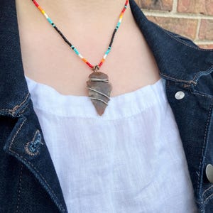 May include: A beaded necklace with a brown arrowhead pendant wrapped in silver wire. The necklace features a colorful beaded strand with black, turquoise, orange, yellow, and red beads.
