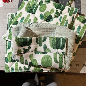 May include: A set of kitchen textiles featuring a cactus print. The set includes two square pot holders with hanging loops, a folded green towel, and a matching oven mitt. The cactus design is in shades of green on a white background.