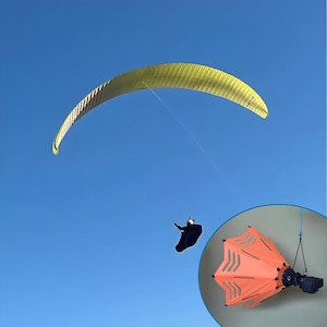 May include: A yellow and black paraglider against a bright blue sky. A person in a black suit is seen beneath the paraglider. An orange, octagonal-shaped object with reflective stripes is also visible.
