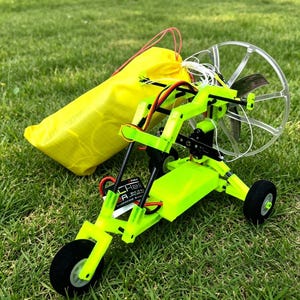 May include: A bright green, three-wheeled paraglider model with a clear propeller guard and a yellow storage bag. The model has black tyres and red and black wiring. The model is on green grass.