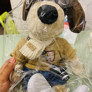 Puede incluir: Un peluche de perro con cuerpo beige, orejas marrones y ojos negros. Viste un suéter beige, una bufanda blanca y vaqueros azules. El juguete está empaquetado en plástico transparente.