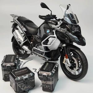 Könnte beinhalten: Ein schwarzes und silbernes BMW R1250 Motorrad mit passenden schwarzen und silbernen Seitenkoffern. Das Motorrad hat eine Windschutzscheibe, einen silbernen Auspuff und das BMW-Logo. Die Seitenkoffer tragen die Buchstaben "GS".