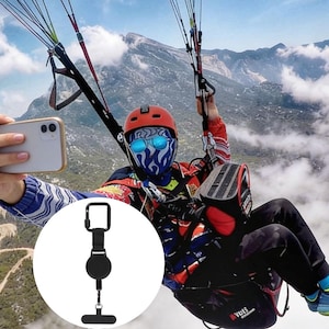 May include: Black retractable phone holder with a carabiner clip and a finger grip. The holder is shown in front of a person paragliding, taking a selfie with a smartphone.